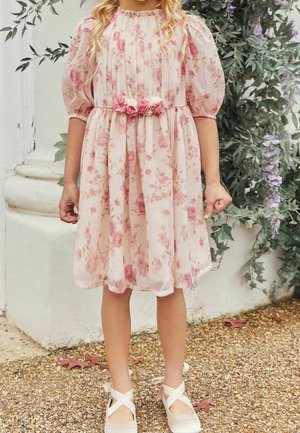 Young girl wearing a pink floral dress with puffed sleeves and flower belt, standing outdoors on gravel near plants and a white wall.