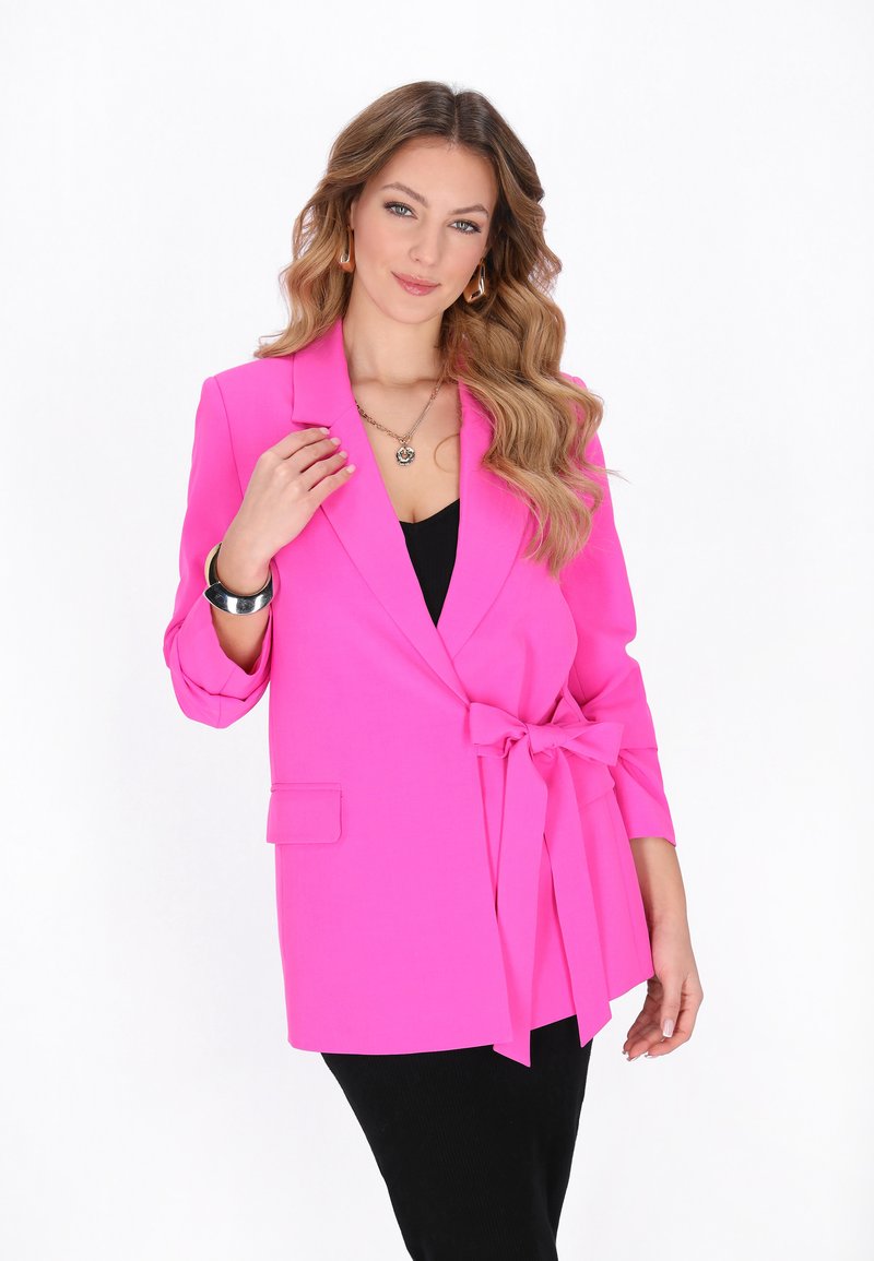 Donna con capelli ondulati indossa un blazer rosa acceso con un fiocco annodato, un vestito nero, un braccialetto d'argento, una collana con ciondolo e orecchini geometrici.