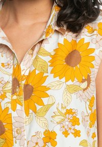 Camisa de botones floral con grandes girasoles amarillos y flores rosas claras sobre un fondo blanco; de manga corta y con un cuello clásico.