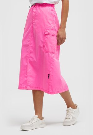 Heller pinker Midirock mit großer seitlicher Tasche, getragen mit weißen Sneakers und weißem Oberteil, an einer Person, die vor einem einfarbigen Hintergrund steht.