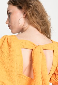 Top amarelo texturizado com mangas bufantes e detalhe de nó nas costas. Grande brinco de argola dourada visível. O fundo é neutro e simples.