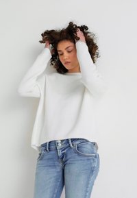White ribbed knit sweater with long sleeves, paired with light blue fitted jeans. The outfit features a relaxed fit and textured fabric.