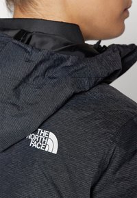 Närbild av en person som bär en mörk, texturerad North Face-jacka med huva, där logotypen syns på baksidan av axeln.