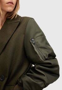 Chaqueta verde oliva con cuerpo de lana y mangas acolchadas y brillantes, con un bolsillo con cremallera en el brazo izquierdo. Cierre de botones y cuello con solapa.