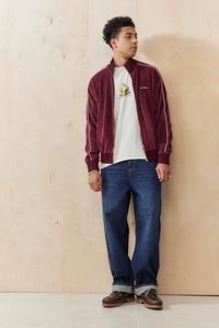 Velvet burgundy jacket with white stripes, worn over a beige graphic T-shirt, paired with loose blue denim jeans and brown shoes.