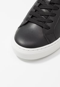 Gros plan sur l'avant d'une chaussure de sport en cuir noir avec des lacets noirs et une semelle épaisse en caoutchouc blanc sur fond blanc.