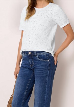 Femme portant un haut blanc à manches courtes en tissu ajouré et un jean bleu en denim, tenant un sac à franges beige sur un fond clair uni.