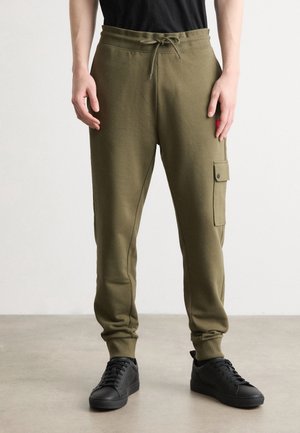 Mann trägt olivgrüne Jogginghose mit Seitentasche, schwarze Sneakers und schwarzes Hemd, steht auf einem hellgrauen Boden.