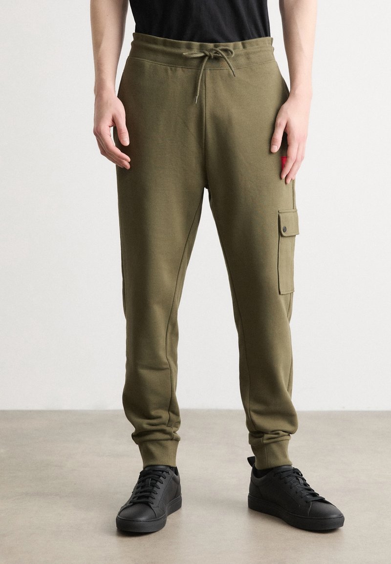 Homme portant un pantalon de jogging vert olive avec poche latérale, baskets noires et chemise noire, debout sur un sol gris clair.