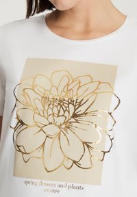 Biała koszulka z krótkim rękawem z dużym złotym wzorem kwiatowym i napisem poniżej "kwiaty i rośliny wiosenne, zał. 1990".