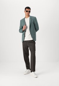 Tirkiz sako, bijela majica, tamne hlače i bijele tenisice. Cijeli izgled karakterizira elegantan dizajn s krojenim fitom i modernim naočalama.