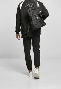 Mochila negra con múltiples bolsillos, correas ajustables y detalle de logo. La persona viste una sudadera y pantalones deportivos negros. Se ven zapatillas blancas.