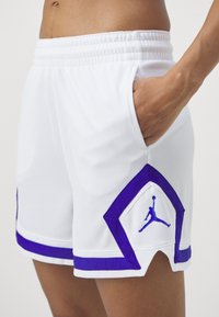 Valged spordilühikesed püksid lilla aktsentidega, millel on võrkpinna tekstuur, küljetaskud ja vasakul küljel Jumpman logo.