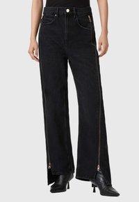 Black denim wide-leg pants with gold hardware, featuring side zippers. The fabric has a slightly textured appearance and a high waist.
