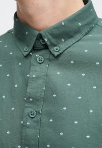 Dunkelgrünes Hemd mit Kragen, verziert mit kleinen blauen und weißen Rautenmuster, passenden Knöpfen und einem weichen, strukturierten Stoff.