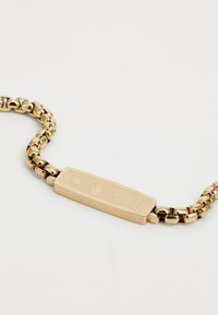 Bracelet en chaîne dorée avec une plaque d'identité rectangulaire ornée de logos en relief et de motifs géométriques ; les maillons polissés créent une texture brillante.