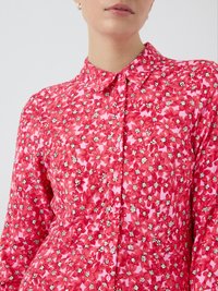 Blouse rouge à fleurs avec un col, une fermeture à boutons à l'avant et des manches longues. Le tissu présente un motif texturé avec des fleurs blanches et roses.