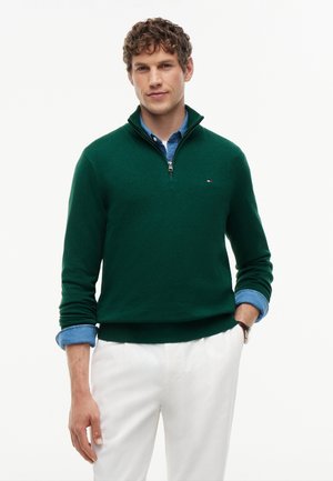 Junger Mann mit lockigem Haar, der einen dunkelgrünen Viertel-Reißverschluss-Pullover über einem blauen Hemd und weißen Hosen trägt, steht mit einer Hand in der Tasche.