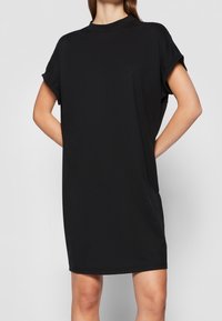 Czarna sukienka T-shirt wykonana z miękkiego materiału, luźny krój z krótkimi, podwiniętymi rękawkami, okrągłym dekoltem i długością do kolan. Gładka struktura i minimalistyczny design.