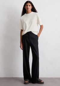 Camicia bianca a maniche corte, vestibilità comoda, abbinata a pantaloni ampi neri. Modello che indossa sandali scuri senza fascetta, in piedi su uno sfondo chiaro.