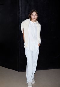 White button-up shirt paired with light blue pants and a cream knitted sweater draped over shoulders. Smooth material and relaxed fit.