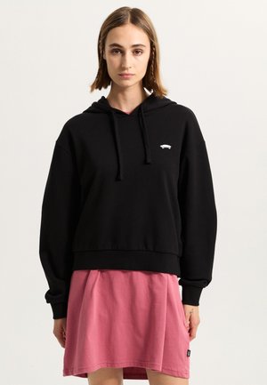 Schwarze, kürzere Kapuzenjacke aus Baumwolle mit einer vorderen Kängurutasche und Kapuze mit Kordelzug, kombiniert mit einem rosa Rock.