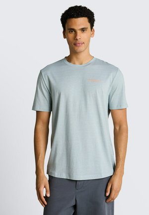 Mężczyzna z kręconymi włosami, ubraną jasnoniebieską koszulkę z krótkim rękawem i okrągłym dekoltem oraz ciemne spodnie, stojący na jednolitym tle.