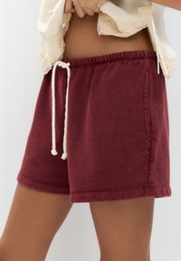 Burgundy sweatshorts med resårmidja, krämfärgad snörning, sidofickor och en mjuk textur. Sytt fåll och logotypdetalj synlig.