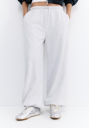 Osoba oblečená v svetlosivých širokých teplákoch s elastickým pásom a strieborných teniskách, stojaca s rukami v vreckách na bielom pozadí.