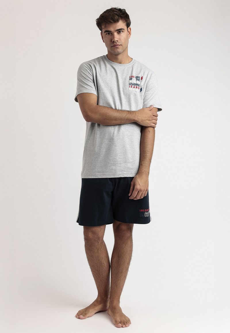 Camiseta gris de manga corta con estampado gráfico y pantalones cortos azules con detalle en texto. El modelo está descalzo, con los brazos cruzados, sobre un fondo neutral.