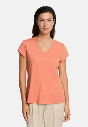 Kobieta w koralowym, krótkim rękawie, T-shircie z dekoltem w serek i jasnobeżowych spodniach, stojąca na tle jednolitego białego tła.