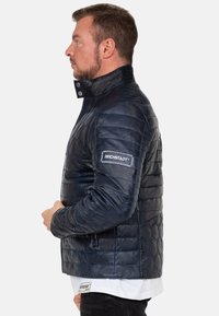 Marineblauwe gewatteerde jas met hexagonaal geborduurd patroon, hoge kraag, zijritsen en een logo patch op de mouw.