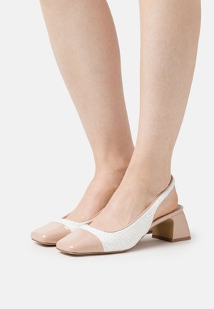 Jalat, joissa valkoiset slingback-lohkokorkokengät beigen lakananilakan varvassuojuksilla, näytetty yksiväristä vaaleaa taustaa vasten.