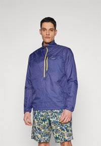 Giacca leggera azzurra con zip gialla, progettata con colletto alto e maniche lunghe. Abbinata a pantaloni corti a fantasia.