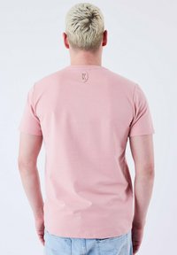 Camiseta corta de manga corta de color rosa claro, hecha de una tela suave, con un escote redondo y un pequeño detalle de logo en la parte de atrás. Diseño simple.