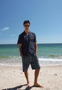 Homme portant des lunettes de soleil, une chemise à manches courtes sombre et un short rayé, debout pieds nus sur une plage de sable avec des vagues de l'océan en arrière-plan.