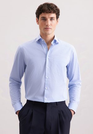 Mann trägt ein hellblaues gemustertes Langarmhemd, das in eine dunkle, gefaltete Hose gesteckt ist, und steht mit den Händen in den Taschen.