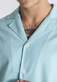 Camicia azzurra chiara con colletto, dotata di due bottoni bianchi. Il tessuto appare liscio con una sottile trama.