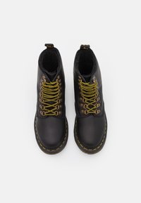 Botas de cuero negro con un forro suave, cordones de color amarillo dorado y ojetes metálicos. Presentan una suela de goma negra con un detalle de costura amarilla.