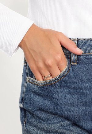 Main avec une bague en forme de nœud en or rose reposant dans la poche avant d’un jean en denim bleu, portant un haut blanc à manches longues côtelé.