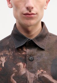Chemise col en tissu marron foncé avec un motif peint. Fermeture à deux boutons au col, avec une surface texturée et des variations de couleur subtiles.