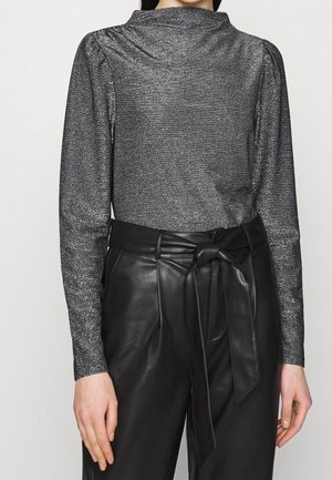 Femme portant un haut à manches longues gris métallisé rentré dans un pantalon en cuir noir taille haute avec une ceinture nouée à la taille.