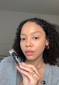 Fond de teint en stick dans un tube transparent avec une base noire. Cheveux bouclés, peau douce. Vernis à ongles bleu clair et accents de bijoux en or.