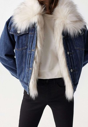 Vrouw die een blauwe spijkerjack draagt met witte nepbontvoering en kraag, over een wit shirt en zwarte broek, staand met handen achter de rug.