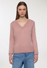 Różowy sweter z dzianiny z dekoltem w serek, prążkowanymi mankietami i dołem oraz luźnym krojem. Nosi się go na wysokich, niebieskich dżinsach.