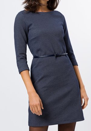 Femme portant une robe ajustée bleu foncé texturée avec des manches trois-quarts et une fine ceinture noire à la taille sur fond blanc.