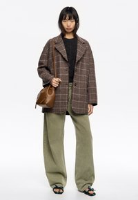 Abrigo de cuadros marrón de gran tamaño, suéter negro, pantalones de pierna ancha color oliva y sandalias multicolores. Bolso cruzado marrón como accesorio.