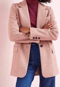 Světle růžový blejzr s vzorem rybí kosti, s klopami, dvěma předními kapsami a černými knoflíky, nošený přes vínový rolák.