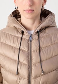 Giacca a vento beige con cappuccio, design a trapuntatura, lacci neri e zip argentata. Texture liscia con una finitura leggermente lucida.