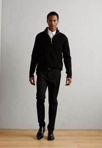 Maglione nero con zip e design testurizzato, indossato sopra una camicia bianca con colletto. Abbinato a pantaloni neri aderenti e scarpe nere lucide.
